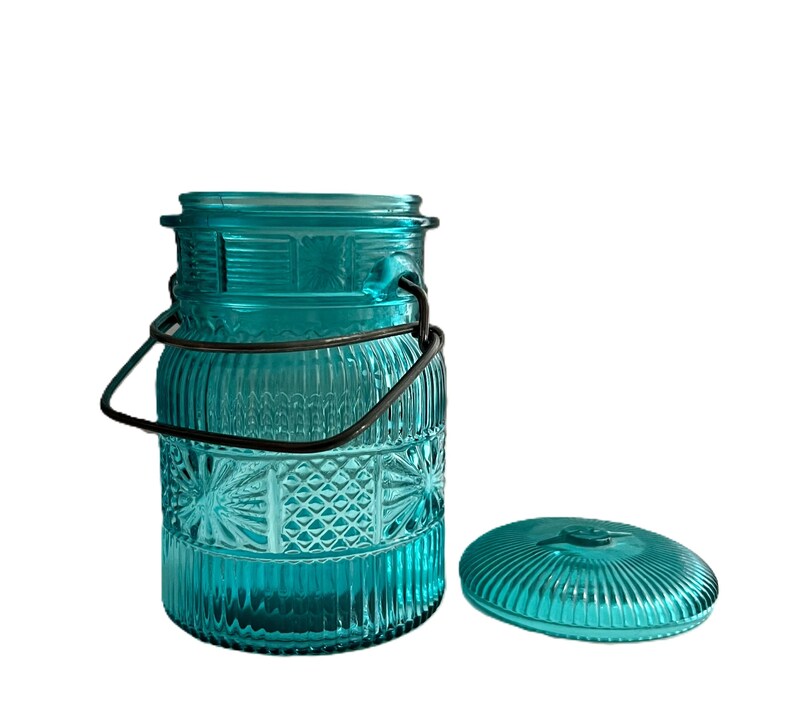 May include: A teal glass jar with a decorative pattern and a metal handle. The jar has a matching lid and is designed with vertical and starburst patterns. The jar is empty and sits on a white background.