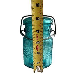 May include: A turquoise glass jar with a lid and a metal handle. The jar is approximately 5 inches tall. The jar has a textured design with vertical lines and diamond patterns. The lid is also textured and has a metal handle.