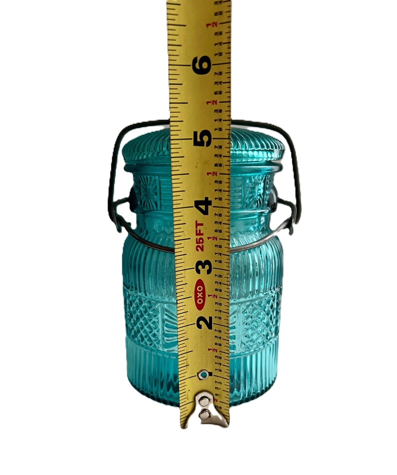 May include: A turquoise glass jar with a lid and a metal handle. The jar is approximately 5 inches tall. The jar has a textured design with vertical lines and diamond patterns. The lid is also textured and has a metal handle.