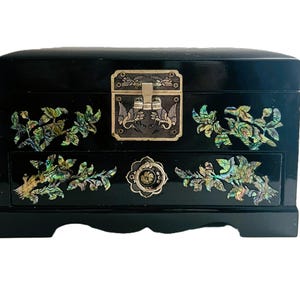 May include: A black jewelry box with a glossy finish, featuring intricate floral and leaf designs inlaid with iridescent abalone shell. The box has a silver-toned clasp and drawer pulls, adding to its ornate aesthetic.
