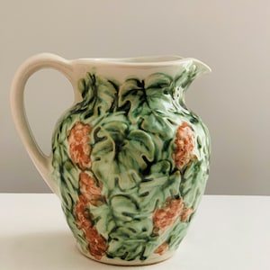 May include: A white ceramic pitcher with a green and pink floral design. The pitcher has a wide mouth and a handle.