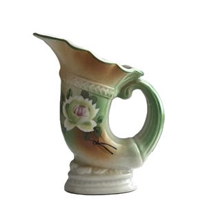 May include: A ceramic pitcher with a curved spout and handle. The pitcher features a floral design with green, white, and pink accents. The body of the pitcher is a light beige color, with a green and brown trim.
