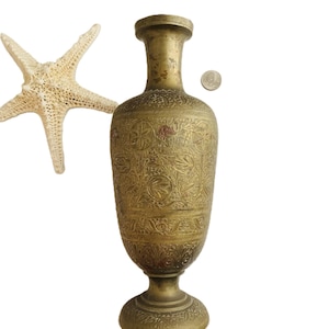 May include: A tall, cylindrical brass vase featuring intricate, hand-etched floral and leaf patterns across its surface. The vase has a narrow neck, a flared rim, and a pedestal base. A white starfish and a small silver coin are placed nearby.