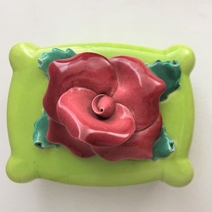 May include: A ceramic wall hanging featuring a large, dimensional red rose with green leaves, set against a bright green, pillow-shaped background. The rose has detailed petals and a glossy finish.