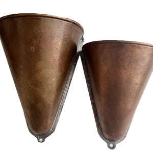 May include: Two cone-shaped copper wall pockets with a metallic finish and a flat back edge. Each piece features a small circular hole at the bottom point for mounting purposes, displaying a weathered patina across the smooth, curved surfaces.