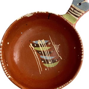 May include: Handmade terracotta bowl with a handle. The bowl is a warm reddish-brown color with white, blue, and green painted accents. The rim has a white dotted pattern. A unique piece for serving or display.