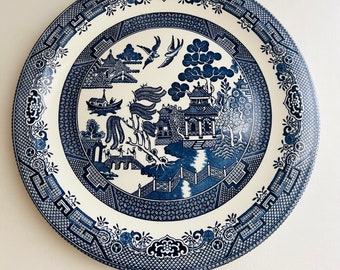 May include: A white ceramic plate with a blue and white willow pattern. The plate features a traditional Chinese landscape scene with a pagoda, trees, and a river.