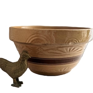 May include: A tan ceramic mixing bowl with a wide dark brown horizontal stripe and embossed arched patterns around the exterior, accompanied by a small, textured brass chicken figurine standing to the left of the bowl.