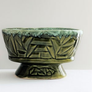 May include: A green ceramic planter with a leafy design and a pedestal base. The planter has a glazed finish and a slightly iridescent green color.