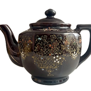 May include: A dark brown ceramic teapot with a lid and handle. The teapot is decorated with gold floral patterns and colorful accents. The spout and handle have a glossy finish, and the teapot sits on a small base.