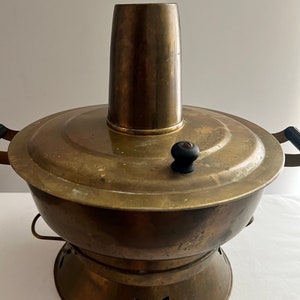 May include: A brass hot pot with a round base and a tall, cylindrical chimney. The pot has two black handles and a black knob on the lid.