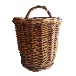 May include: A woven brown basket with a handle. The basket is made of natural materials and has a cylindrical shape with a slightly flared top. The handle is made of twisted material.