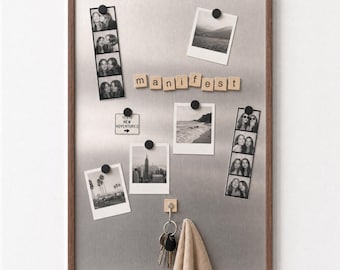 May include: A rectangular magnetic bulletin board with a dark wood frame and a metallic silver surface. It features black circular magnets holding black and white photographs, a small sign reading "YES TO NEW ADVENTURES," and wooden letter tiles spelling "manifest." A hook holds a set of keys and a beige cloth.