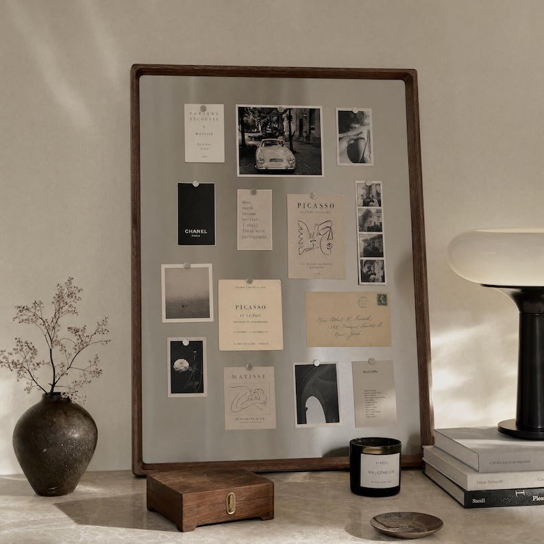 A large stainless steel magnetic board with a dark walnut wood frame stands on a table, holding various photos and notes attached with small magnets, flanked by a dark ceramic vase, small wooden box, and candle.