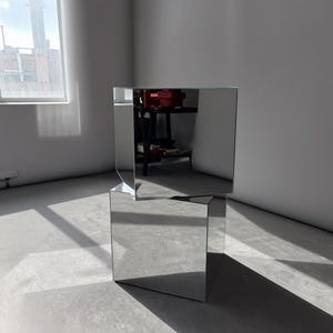May include: A modern, mirrored side table with a unique geometric design. The table is constructed from reflective glass panels, creating a cube-like shape with an offset top. The design reflects the surrounding environment, adding a contemporary touch to any space.