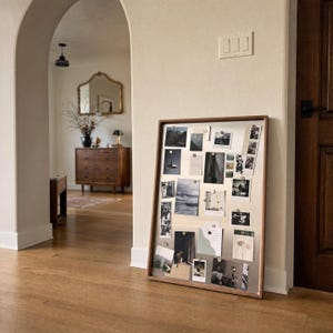 Large magnetic board leaning against a beige wall on a wooden floor, densely covered with various photos and notes held by small magnets.