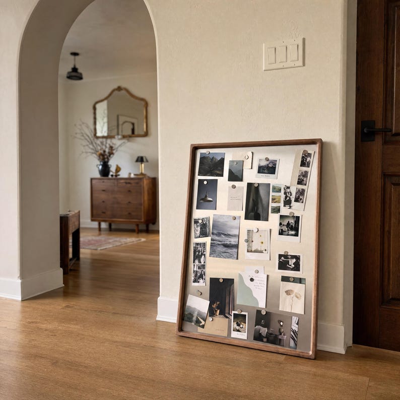 Large magnetic board leaning against a beige wall on a wooden floor, densely covered with various photos and notes held by small magnets.