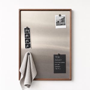 Walnut-framed magnetic board mounted on a white wall, holding two photos, a black to-do list, and a hanging grey hand towel.