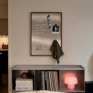 Magnetic board hanging on a wall above a metal shelf holding records and a pink lamp, with a to-do list, photos, sticker, and olive towel attached.