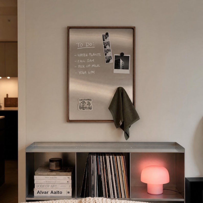 Magnetic board hanging on a wall above a metal shelf holding records and a pink lamp, with a to-do list, photos, sticker, and olive towel attached.