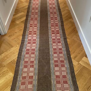 Puede incluir: Una alfombra pasillera larga y rectangular con un panel central de color marrón oscuro flanqueado por dos bandas anchas de patrón a cuadros rojo y crema. Los bordes tienen rayas finas azules y blancas y flecos en los extremos.