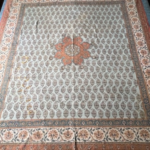 Op de afbeelding: Grote, rechthoekige textiel met een lichtblauwe achtergrond en een herhalend patroon van kleine, donkerbruine bloemmotieven. Het midden heeft een groot, oranje bloemmotief. De rand heeft een bloemen- en geometrisch patroon in bruin- en oranje tinten.