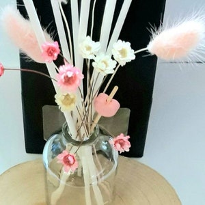 May include: A clear glass bottle with white diffuser sticks and pink and white dried flowers. The bottle is sitting on a wooden surface.