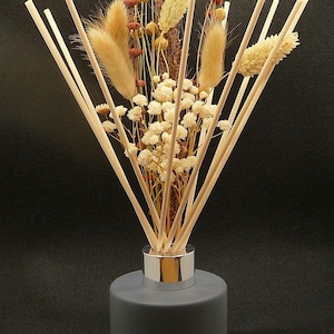May include: A grey reed diffuser with a silver collar, filled with dried flowers and reeds. The arrangement includes beige, brown, and white dried flowers and light-coloured reeds. The diffuser sits on a wooden base against a dark background.