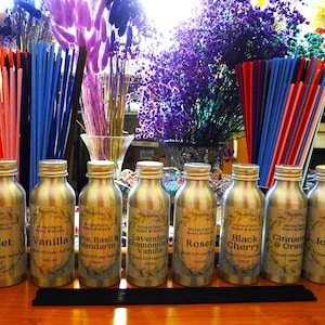 May include: A selection of reed diffuser refills in silver metal bottles, each labelled with a different scent: Violet, Vanilla, Lime Basil & Mandarin, Lavender Chamomile Vanilla, Roses, Black Cherry, Cinnamon & Orange, and Ice Pine. The background features colourful dried flowers and reeds.