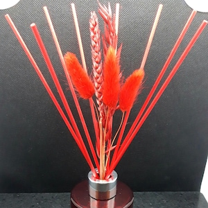 May include: Red diffuser sticks in a glass bottle with a silver cap. The sticks are surrounded by red dried flowers and wheat.