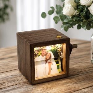 May include: A square dark wood photo display box with an internal light illuminating a photograph of a couple in wedding attire. The box features a small gold button on the top surface and a wooden crank handle on the side for rotating the image.