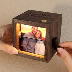 May include: A dark wooden photo cube with a picture of a couple illuminated inside. The top of the cube is engraved with the words "Together Wherever". A small gold button is on top, and a black knob is on the side.