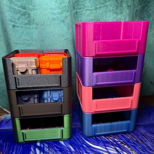 May include: Two stacks of colorful, 3-tiered storage containers. The left stack has a black frame with green, brown, and black drawers. The right stack has a pink, purple, coral, and blue color scheme. Each drawer is designed to hold small items.