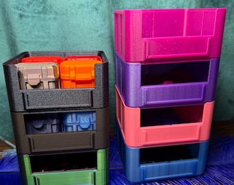 May include: Two stacks of colorful, 3-tiered storage containers. The left stack has a black frame with green, brown, and black drawers. The right stack has a pink, purple, coral, and blue color scheme. Each drawer is designed to hold small items.