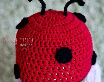 May include: A red crocheted ladybug hat with black spots and antennae. The hat is worn by a child with purple clothing.