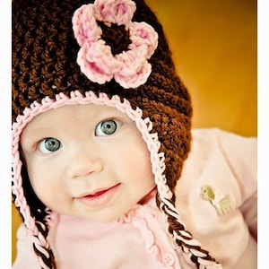 May include: A baby wearing a brown crocheted hat with a pink flower on top. The hat has brown and pink braided ties. The baby is wearing a pink sweater with white buttons and ruffles.