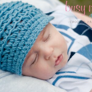 Op de afbeelding: Een baby draagt ​​een blauwe gehaakte muts met een rand. De muts heeft een textuurpatroon. De baby slaapt en draagt ​​een wit en blauw gepatroneerd shirt.