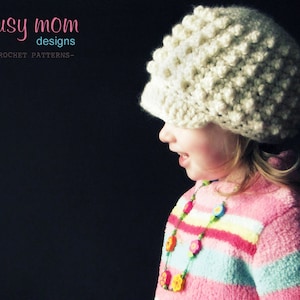 Peut inclure: Une jeune personne portant un bonnet en crochet blanc avec un design texturé. Le bonnet est tiré vers le bas sur le front et couvre les oreilles. Elle porte un pull rayé rose et blanc avec un collier de perles colorées.