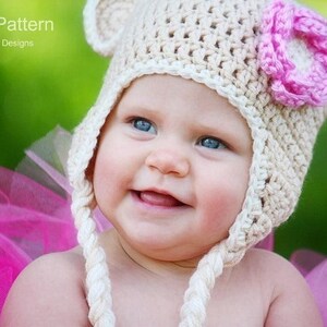 May include: A baby wearing a crocheted bear hat with a pink flower on top. The hat is cream colored with white trim. The baby is wearing a pink tutu.