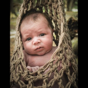 May include: A baby is nestled in a crocheted hammock made of beige yarn. The hammock is a simple design with a repeating pattern of knots. The baby is looking directly at the camera with a serious expression.