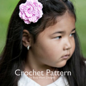 May include: A young girl with long dark hair wearing a white collared shirt with a pink crocheted flower hair clip. Crochet Pattern by Busy Mom Designs.