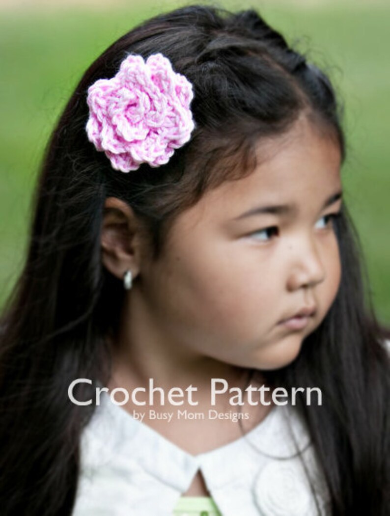 May include: A young girl with long dark hair wearing a white collared shirt with a pink crocheted flower hair clip. Crochet Pattern by Busy Mom Designs.