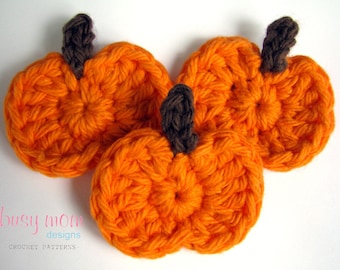 May include: Three crocheted orange pumpkins with brown stems. The pumpkins are arranged in a cluster on a white background.