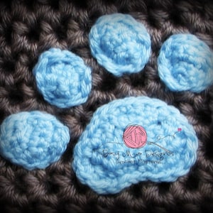 May include: Close-up of a crocheted paw print with five blue circles. The paw print is on a brown crocheted background. The text "Busy Mom Designs Crochet Patterns" is visible in the center of the paw print.