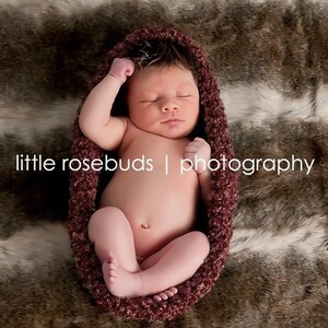 May include: A newborn baby is sleeping peacefully in a burgundy crocheted swaddle on a brown and white faux fur blanket. The baby is wearing a neutral expression and has their eyes closed.