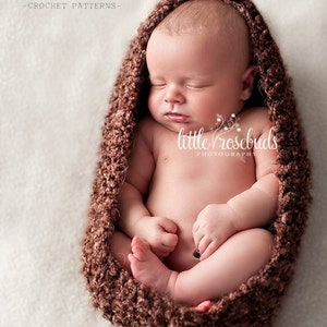 May include: A newborn baby sleeps peacefully inside a brown crocheted cocoon. The baby is nestled in a soft, textured, brown cocoon, with the text "busy mom designs - crochet patterns" and "little rosebuds photography" visible.