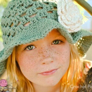 May include: A young girl wearing a green crochet sun hat with a white crochet flower embellishment. The hat has a wide brim and a crocheted design. The girl has freckles and is looking at the camera.