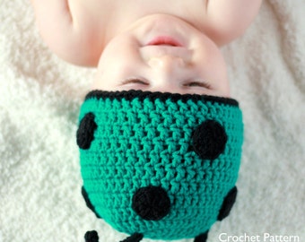 May include: A green crocheted ladybug hat with black spots and antennae. The hat is worn by a baby lying on a white blanket.