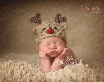 May include: A newborn baby wearing a crocheted reindeer hat with a red nose, sleeping on a white fluffy blanket. The baby is wearing a brown crocheted hat with a red nose and antlers. The baby is sleeping on a white fluffy blanket. The baby is wearing a brown crocheted hat with a red nose and antlers. The baby is sleeping on a white fluffy blanket.