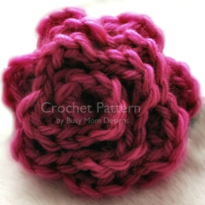 May include: A close-up of a crocheted rose made with a deep pink yarn. The rose is made with a simple stitch pattern and has a textured look.
